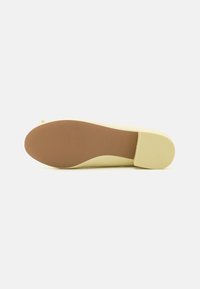 Zapato amarillo de tacón bajo con una parte superior lisa, suela de goma ligeramente texturizada y punta redondeada. Presenta un tacón modesto y un detalle de marca sutil.