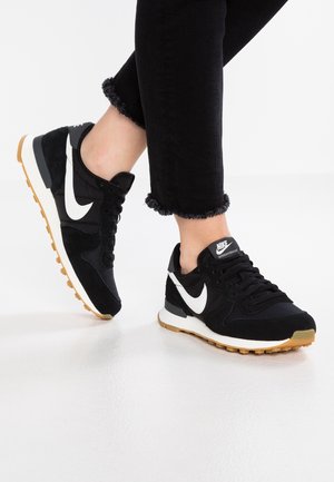 Personne portant des baskets Nike noires avec un swoosh blanc et des semelles en gomme, assorties à un pantalon noir à ourlet effiloché sur un sol blanc.