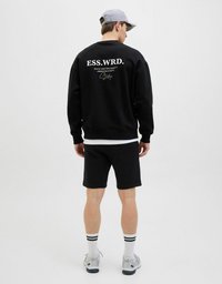 Czarny sweatshirt z białym napisem na plecach, w połączeniu z czarnymi szortami, białymi skarpetkami z czarnymi paskami i jasnoszarymi trampkami.