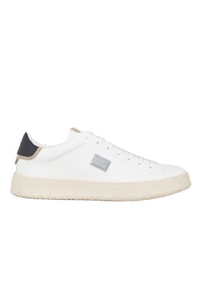 Zapatilla baja blanca con cordones en la parte frontal, suela texturizada color blanco roto, lengüeta negra en el talón y una pequeña placa de logo plateada en el lateral.