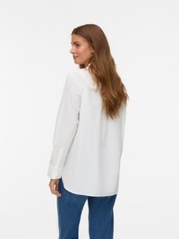 White long-sleeve shirt with a collar, featuring a smooth texture and slightly curved hem, paired with blue denim pants.