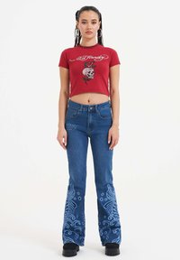 Rød kortærmet T-shirt med broderet kranium og rosedesign, parret med flagrende blå jeans med detaljeret blå broderi på nederste ben.