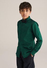 Zelena košulja s gumbima i uzorkom s točkicama, koja ima klasičan ovratnik i duge rukave. Uparena s crnim leptir-mašnom. Crne hlače.