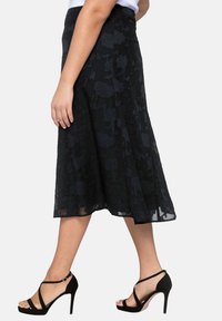 Mujer vestida con una falda midi negra con flores y sandalias de tacón negro con tiras en el tobillo, de pie contra un fondo blanco.