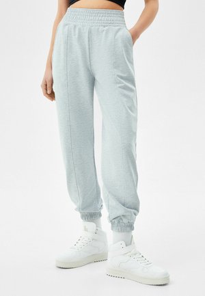 Person, die hellgraue Jogginghosen und weiße High-Top-Sneaker trägt, steht mit den Händen in den Taschen vor einem einfarbigen weißen Hintergrund.