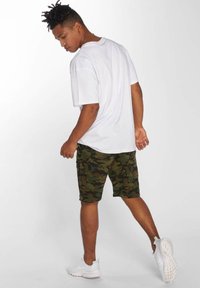 T-shirt bianco a maniche corte abbinato a pantaloni corti camouflage verdi e scarpe da ginnastica bianche; il modello è visto da dietro con uno sfondo neutro.