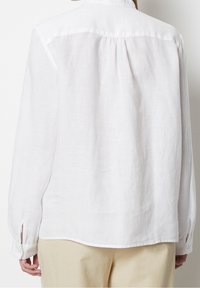 Chemise en lin blanche avec une coupe décontractée, des poignets à boutons et un pli au dos. Le tissu présente un aspect texturé et respirant.