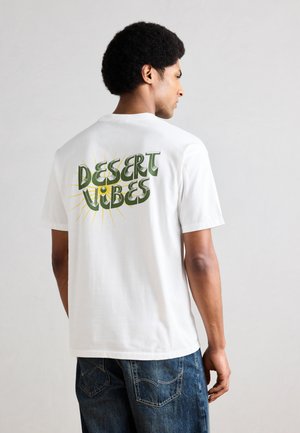 Mann iført hvit T-skjorte med grønn tekst «DESERT VIBES» og gule solstråler på baksiden, kombinert med blå jeans, mot ensfarget bakgrunn.