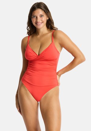Woman with shoulder-length brown hair smiling, wearing a red one-piece swimsuit with thin shoulder straps against a plain background.