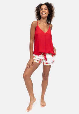 Femme souriante aux cheveux bouclés, debout pieds nus, portant un haut rouge sans manches et un short blanc à fleurs, les mains dans les poches, sur fond blanc.