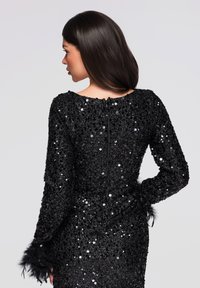 Robe noire en sequins à manches longues, avec des poignets ornés de plumes et une fermeture éclair au dos. Le modèle est cintré avec un col rond.