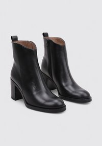 Botas negras de cuero hasta el tobillo con un alto tacón cuadrado, cremallera lateral, textura suave y punta redondeada. El interior presenta un forro marrón claro.