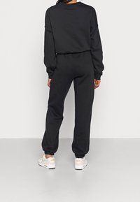 Black cotton jumpsuit with long sleeves and elastic cuffs, featuring a relaxed fit. Paired with white sneakers with orange accents.