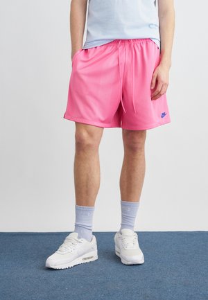 Persona que lleva pantalones cortos Nike de color rosa brillante, calcetas azul claro y zapatillas blancas, de pie sobre un suelo alfombrado azul frente a una pared lisa.