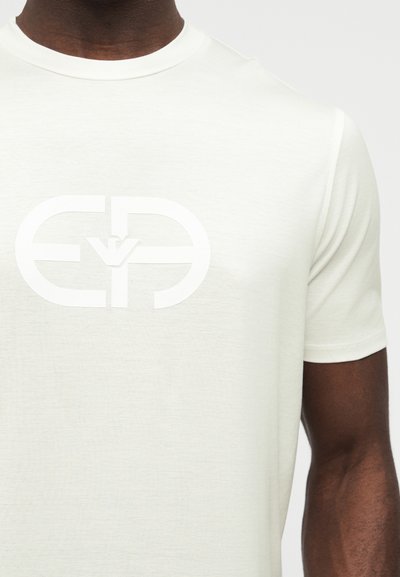 Κοντινό πλάνο ενός ατόμου που φοράει λευκό μπλουζάκι με μεγάλο λευκό λογότυπο Giorgio Armani τυπωμένο στο στήθος.