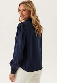 Donna con lunghi capelli biondi che indossa una blusa a maniche lunghe blu navy e pantaloni bianchi, sorridente e che guarda oltre la spalla.