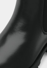 Gros plan sur une botte Chelsea en cuir noir lisse avec panneau latéral élastique et petit talon sur fond blanc.