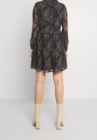 Svart klänning med lila och gröna blommönster, långa ärmar, utsvängd kjol och dragkedja bak. Modellen bär beiga klackstövlar med blockklack.