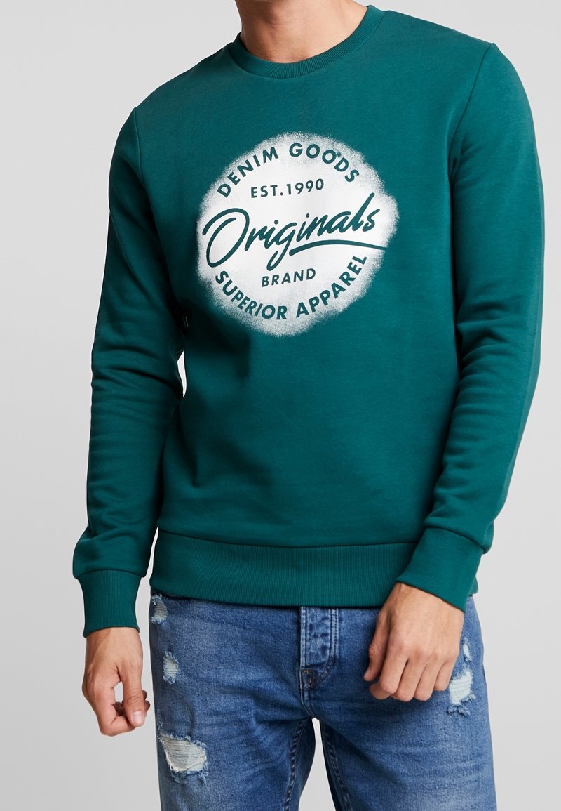 Homme portant un sweat-shirt turquoise avec un logo circulaire blanc indiquant « Denim Goods, Originals, Superior Apparel, Est. 1990 » et un jean bleu déchiré.