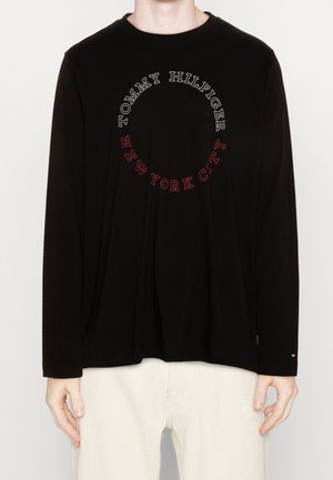 Osoba nosząca czarną koszulkę z długim rękawem z napisem "Tommy Hilfiger New York City" w okręgu na piersi oraz jasne spodnie.