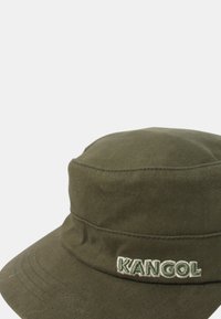 Gorra de cubo de color verde oliva hecha de algodón. Cuenta con un ala ancha, detalles de costura y un parche logo en la parte delantera con "KANGOL" en verde claro.
