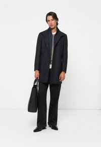 Homme debout contre un fond blanc, portant un manteau noir, un pull à motif, un pantalon noir, des chaussures noires et tenant un sac fourre-tout noir dans une main.