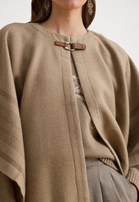 Donna che indossa un cappotto di lana beige con chiusura a fibbia in pelle, un dolcevita, pantaloni grigi plissettati e orecchini di cristallo pendenti.