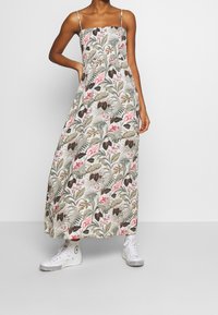 Robe longue à fines bretelles, motif floral avec feuilles vertes et fleurs roses, en tissu léger. Associée à des baskets blanches.