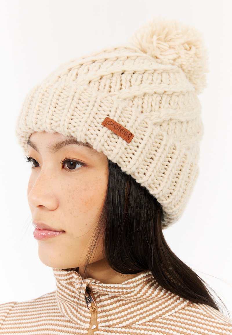 Joven mujer que lleva un gorro de punto color crema con pompón y un suéter beige a rayas con cremallera, mirando hacia la izquierda.