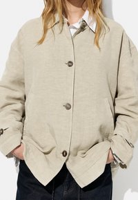 Beige, oversized jakke med en tekstureret linnedfinish. Har rund krave, knaplukning og sidelommer, båret over en hvid skjorte.