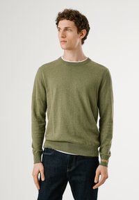 Suéter verde de punto con cuello redondo y mangas largas. Presenta puños y dobladillo acanalados, con un detalle de logo en el puño izquierdo.