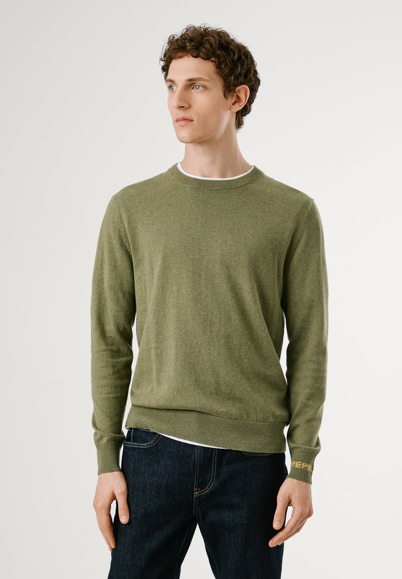 Suéter verde de punto con cuello redondo y mangas largas. Presenta puños y dobladillo acanalados, con un detalle de logo en el puño izquierdo.