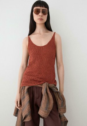 Femme aux longs cheveux foncés et lunettes de soleil portant un débardeur texturé couleur rouille, un pantalon marron et une chemise rayée nouée autour de la taille.