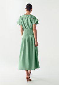 Vestido midi verde con mangas cortas tipo mariposa, cintura ceñida y falda fluida, fabricado en tejido ligero. La vista trasera muestra los pliegues.