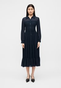 Φόρεμα πουκάμισο μακρυμάνικο σε ναυτικό μπλε, με κουμπιά μπροστά, σφιχτή εφαρμογή στο μπούστο και φούστα σε στρώσεις. Διαφανής υφή με ρέουσα σιλουέτα.