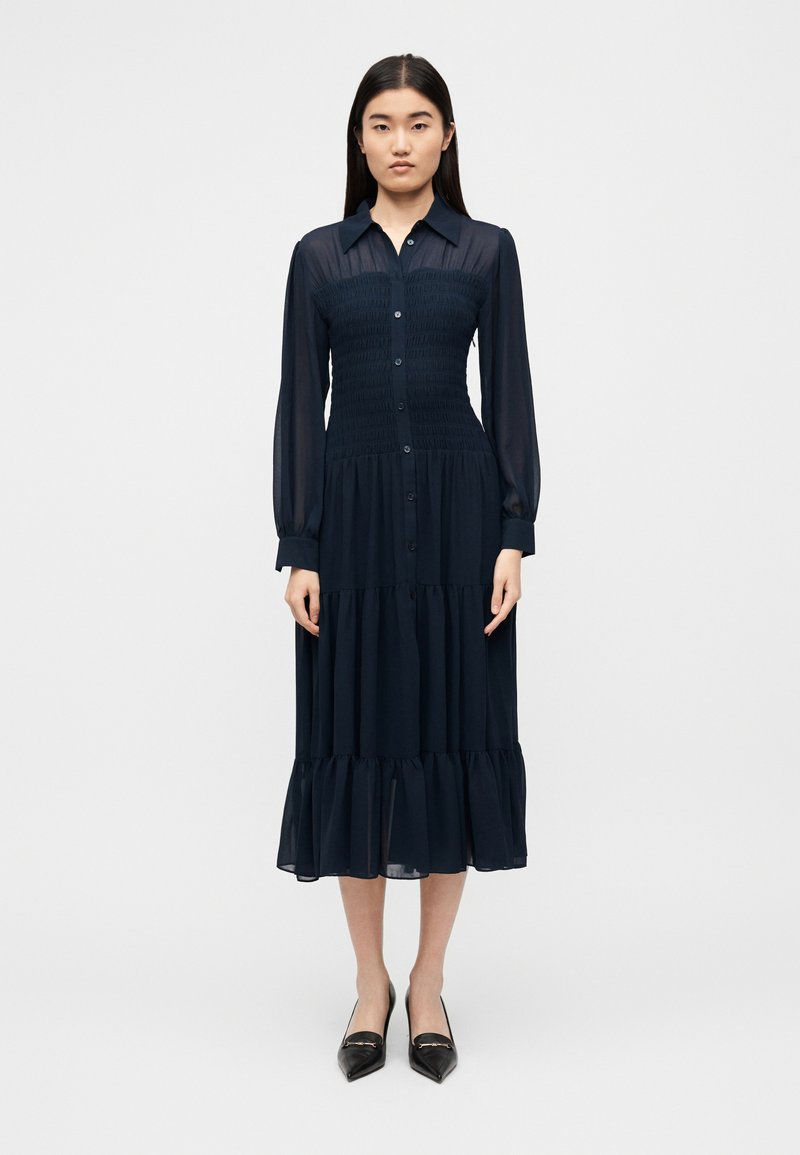 Φόρεμα πουκάμισο μακρυμάνικο σε ναυτικό μπλε, με κουμπιά μπροστά, σφιχτή εφαρμογή στο μπούστο και φούστα σε στρώσεις. Διαφανής υφή με ρέουσα σιλουέτα.