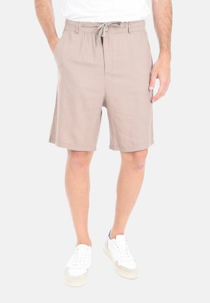 Uomo che indossa pantaloncini beige con coulisse e scarpe da ginnastica bianche, in piedi con le mani in tasca su uno sfondo semplice.
