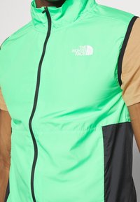 Chaleco verde sin mangas con acentos negros y un cuello alto. Presenta el logo "The North Face" en blanco. Hecho de un material ligero y suave.
