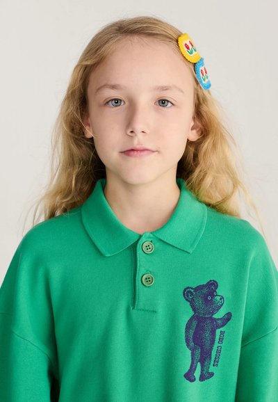 Jeune fille aux longs cheveux blonds porte un polo vert avec un motif d'ours en peluche sombre et des barrettes colorées sur le côté droit.