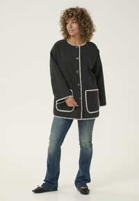 Grå ulljacka med rund halsringning, vit zigzag-kant, stora knappar och framfickor, bärs över utsvängda jeans.