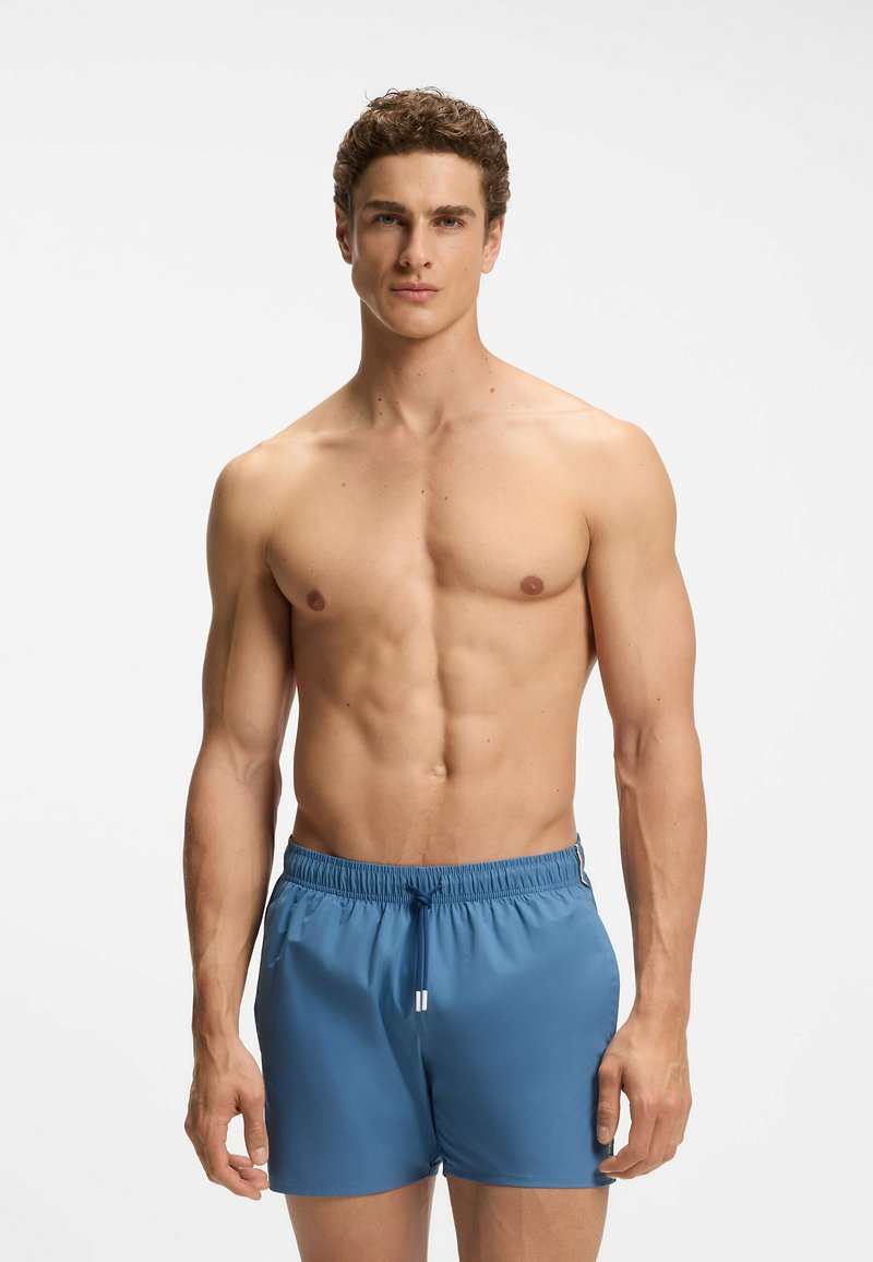 Homme musclé aux cheveux bouclés portant un short de bain bleu, debout devant un fond blanc uni, regardant droit devant.