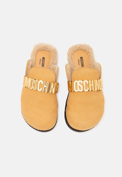 Chaussons beige à enfiler avec doublure duveteuse et semelles noires, avec des lettres dorées formant "MOSCHINO" sur les brides supérieures.