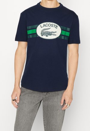 Camiseta Lacoste azul marino con rayas verdes y logo ovalado blanco con un cocodrilo, combinada con vaqueros grises y anillos plateados en los dedos.