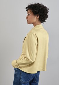 Giacca gialla, strutturata, con colletto e dettagli ai bottoni, indossata sopra pantaloni in denim blu. La silhouette è rilassata e leggermente accorciata.