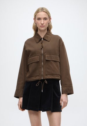 Kobieta o blond włosach w brązowej kurtce zapinanej na zamek z dużymi przednimi kieszeniami i czarnej plisowanej spódnicy na białym tle.