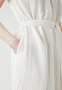 Rochie albă, texturată, cu deschidere cu nasturi în față, cordon la talie și buzunare pe lateral. Material ușor cu un aspect șifonat, care oferă o croială fluidă.