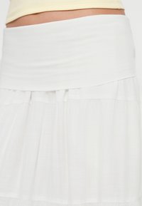 Λευκή φούστα με φαρδιά, μαλακή μέση. Το ύφασμα είναι ελαφρύ και ρέον, με λεπτή υφή και πιέτες κοντά στο στρίφωμα.