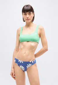 Γυναίκα που φοράει πράσινο πλεκτό κοντό τοπ και μπλε μαγιό με μεγάλα λευκά σχέδια μαργαρίτας, στέκεται μπροστά σε ανοιχτό γκρι φόντο.