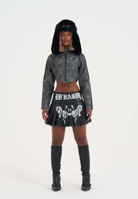 Cropped, gray faux leather jacket with zipper; black pleated skirt featuring "Ed Hardy" text and graphic print. Black fur hat, knee-high boots.