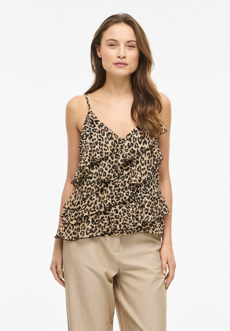 Femme portant un haut sans manches à volants à imprimé léopard avec un pantalon beige, debout devant un fond blanc uni.